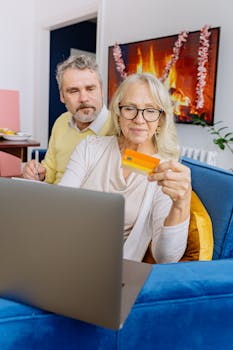 Elderly couple smiles as they shop online with a credit card on a cozy couch.